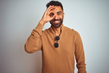 Hint adam zarif kazak ve güneş gözlüğü ayakta izole beyaz arka plan ile el gülümseyen ok jest yapıyor giymiş, göz parmak mutlu yüz ile görünümlü.
