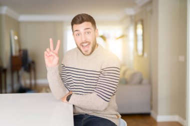 Evde bir kazak giyen genç yakışıklı adam mutlu yüz zafer işareti yapıyor kamera göz kırpıyor ile gülümsüyor. İki numara.