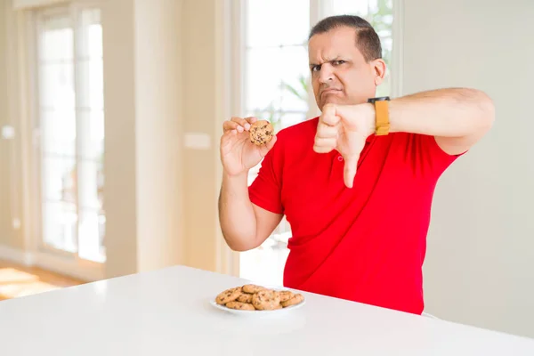 中年男子吃巧克力饼干饼干在家里与愤怒的脸 负面的迹象显示不喜欢与拇指向下 拒绝概念 — 图库照片