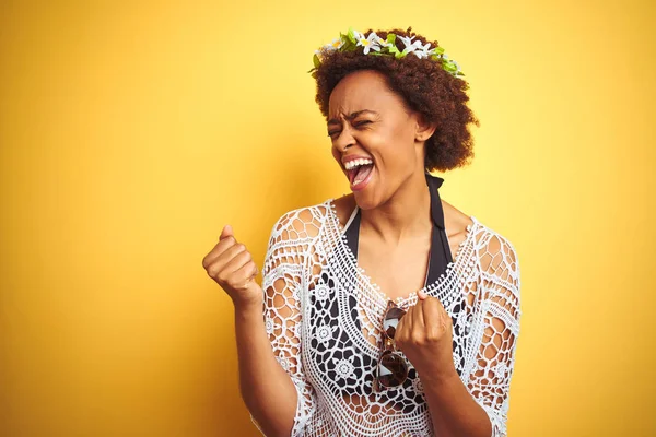 Young african american woman with afro hair wearing flowers crown over yellow isolated background very happy and excited doing winner gesture with arms raised, smiling and screaming for success. Celebration concept.