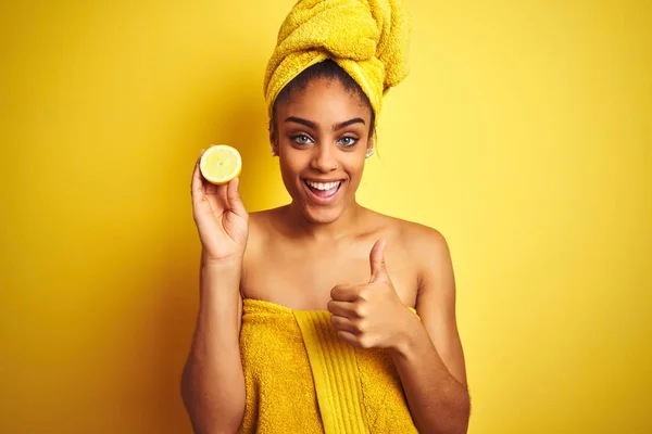 シャワーの後にタオルを着たアフロ女性は 大きな笑顔で幸せな孤立した黄色の背景の上にスライスレモンを保持し 指で親指アップ 優れた兆候 — ストック写真