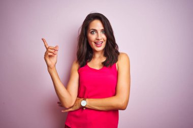 Yüze büyük bir gülümseme ile pembe izole arka plan üzerinde ayakta t-shirt giyen genç güzel kadın, kamera bakarak yan el ve parmak ile işaret.