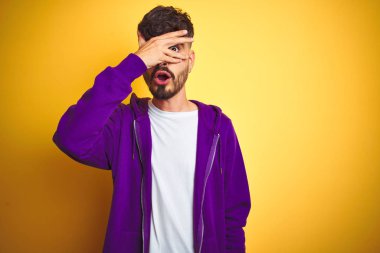 Dövme ile genç adam izole sarı arka plan üzerine spor mor sweatshirt giyen şok kapsayan yüz ve gözleri elle, utanmış ifade ile parmaklarla bakarak.