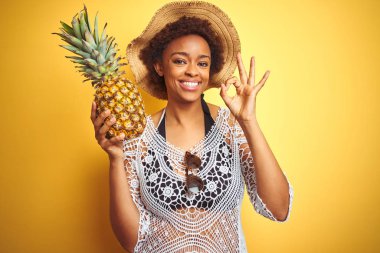 Genç African Amerikan kadın ile afro saç tutan Pineaple üzerinde sarı izole arka plan yapıyor ok işareti ile parmaklar, mükemmel sembol