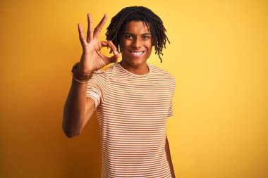 Kıvırcık sarı arka plan üzerinde ayakta çizgili t-shirt giyen dreadlocks ile Afro adam el ve parmaklar ile ok işareti yapıyor pozitif gülümseyen. Başarılı ifade.