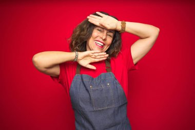 Kırmızı izole arka plan üzerinde önlük üniforma giyen orta yaş yaşlı kadın Gülümseyen neşeli oynarken yüzleri gösteren elleri ile bir boo gözetleme. Şaşırttı ve çıktı