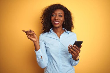 Genç afro-amerikan kadın izole sarı arka plan üzerinde duran akıllı telefon kullanarak çok mutlu yan el ve parmak ile işaret
