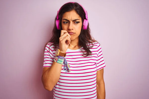 Young beautiful woman listening to music using headphones over isolated pink background looking stressed and nervous with hands on mouth biting nails. Anxiety problem.