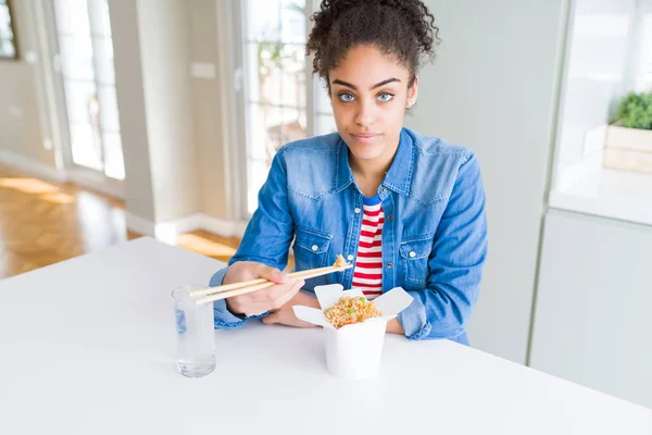 Young african american woman eating asian noodles from delivery box with a confident expression on smart face thinking serious
