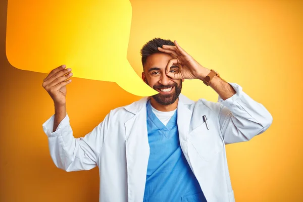 Young indian doctor man holding speech bubble standing over isolated yellow background with happy face smiling doing ok sign with hand on eye looking through fingers