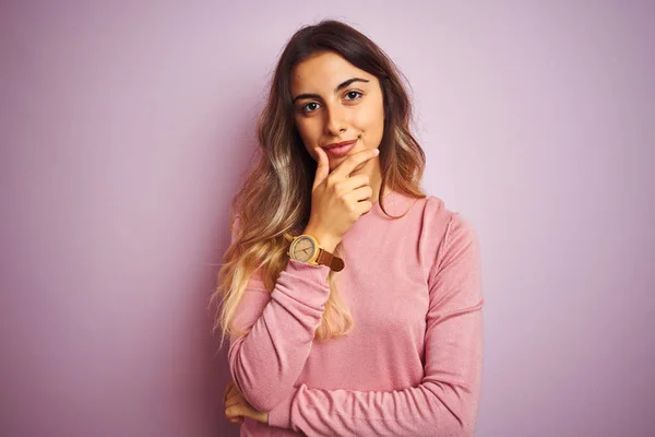 Young beautiful woman wearing a sweater over pink isolated background looking confident at the camera with smile with crossed arms and hand raised on chin. Thinking positive.