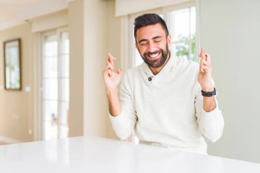 Evde rahat beyaz kazak giyen yakışıklı İspanyol adam umut ve gözler kapalı parmaklar geçiş gülümseyen. Şans ve batıl fikir.