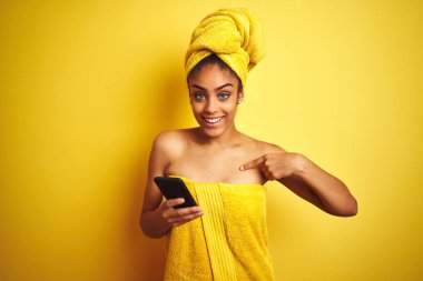Afro kadın havlu giyen duş sonra sürpriz yüz kendini işaret parmak ile izole sarı arka plan üzerinde smatrphone kullanarak