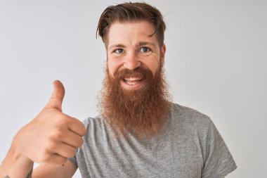 Genç kızıl İrlandalı adam büyük bir gülümseme ok işareti yapıyor mutlu izole gri arka plan üzerinde duran t-shirt giyen, parmakları ile başparmak, mükemmel işareti