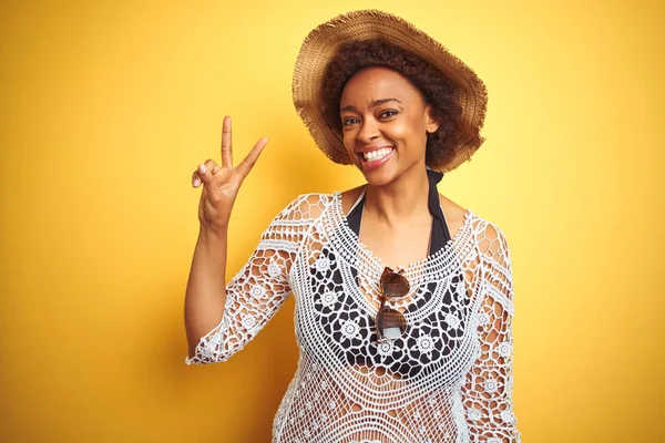 Young african american woman with afro hair wearing summer hat over white isolated background smiling with happy face winking at the camera doing victory sign. Number two.