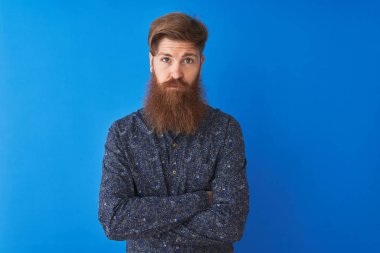 İzole mavi arka plan üzerinde ayakta çiçek Yaz gömlek giyen genç Kızıl İrlandalı adam şüpheci ve sinir, Çapraz kollar ile yüz üzerinde ifade onaylamaz. Negatif kişi.