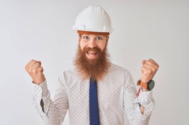 Genç Redhead İrlandalı mimar adam izole beyaz arka plan üzerinde güvenlik kask giyiyor şaşırttı ve silah yükseltilmiş ve açık gözleri ile başarı için hayran kutluyor. Kazanan kavramı.