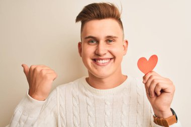 Genç yakışıklı romantik adam izole beyaz arka plan üzerinde duran kağıt kalpleri tutan işaret ve mutlu yüzü gülümseyerek yan başparmak ile gösteren