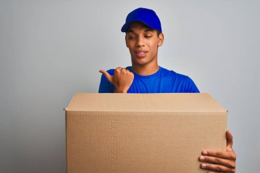 Genç yakışıklı arap teslimat adam izole beyaz arka plan üzerinde duran bir kutu tutan işaret ve mutlu yüzü gülümseyerek yan başparmak ile gösteren