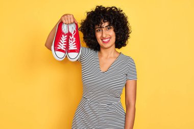 Kıvırcık saçlı genç arap kadın mutlu bir yüz ayakta ve dişleri gösteren kendinden emin bir gülümseme ile gülümseyerek izole sarı arka plan üzerinde rahat spor ayakkabı tutan