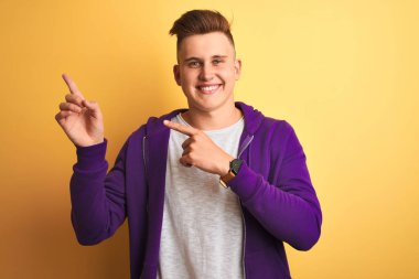 Genç yakışıklı adam izole sarı arka plan üzerinde duran mor sweatshirt giyen gülümseyerek ve yan iki el ve parmakları ile işaret kamera bakarak.