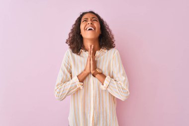 İzole pembe arka plan üzerinde ayakta çizgili gömlek giyen genç Brezilyalı kadın yalvarıyor ve yüz üzerinde umut ifade ile birlikte eller ile dua çok duygusal ve endişeli. Affedilmesini istiyorum. Din konsepti.