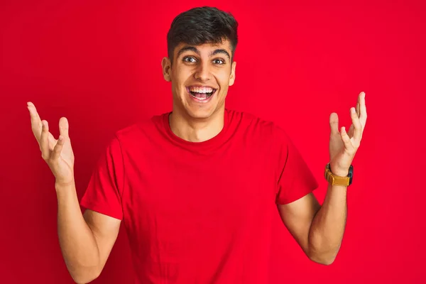 孤立した赤い背景の上に立つTシャツを着た若いインド人男性は 腕を上げて目を開けて興奮して成功に驚きました 勝者のコンセプト — ストック写真