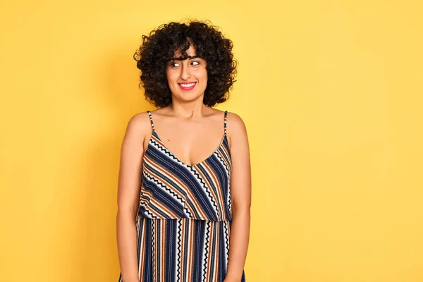 Arab woman with curly hair wearing striped colorful dress over isolated yellow background smiling looking to the side and staring away thinking.