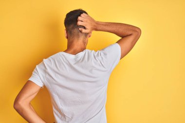 Beyaz tişört giyen genç hintli adam izole sarı arka plan üzerinde duran geriye baş el ile şüphe hakkında düşünme