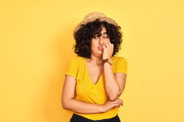 Kıvırcık saçlı genç arap kadın izole sarı arka plan üzerinde t-shirt ve şapka giyen stresli ve ağız tırnak ısırma elleri ile sinirli görünümlü. Anksiyete sorunu.