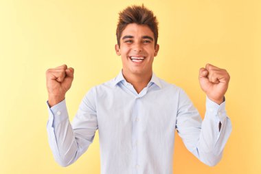 İzole sarı arka plan üzerinde zarif gömlek giyen genç yakışıklı işadamı kollarını kaldırdı ve gözleri açık başarı için sürpriz ve şaşırttı. Kazanan kavramı.