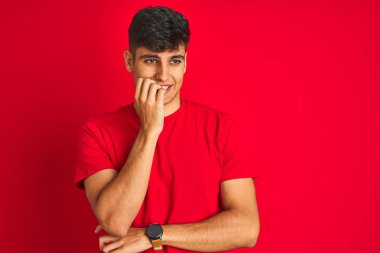 İzole kırmızı arka plan üzerinde duran t-shirt giyen genç hintli adam stresli ve ağız tırnakısırma elleri ile sinirli görünümlü. Anksiyete sorunu.