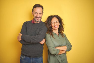 İzole sarı arka plan üzerinde güzel orta yaş çift kamera bakarak çapraz silah ile gülümseyen mutlu yüz. Pozitif kişi.