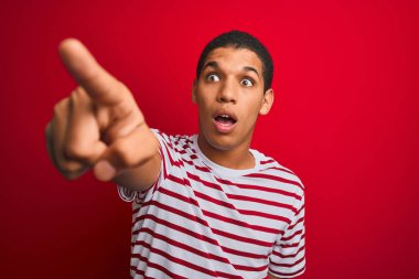 Genç yakışıklı arap adam izole kırmızı arka plan üzerinde çizgili t-shirt giyen parmak ile işaret önünde şaşırttı, açık ağız şaşkın ifade, ön bir şey