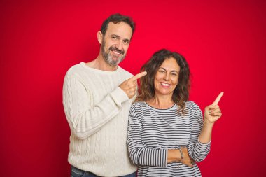 Yüzünde büyük bir gülümseme ile izole kırmızı arka plan üzerinde kış kazak giyen güzel orta yaş çift, kamera bakarak yan el ve parmak ile işaret.