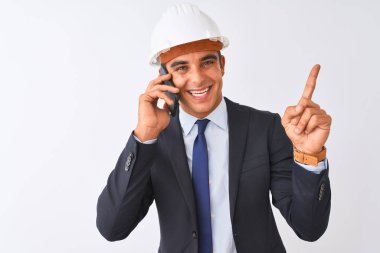 İzole beyaz arka plan üzerinde akıllı telefonda konuşurken kask giyen genç mimar adam çok mutlu yan el ve parmak ile işaret