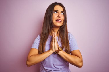 Genç güzel kadın rahat t-shirt izole pembe arka plan üzerinde ayakta giyiyor yalvarıyor ve el ile birlikte umut ifade yüz çok duygusal ve endişeli ile dua. Affedilmesini istiyorum. Din konsepti.