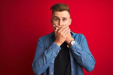 İzole kırmızı arka plan üzerinde duran denim gömlek giyen genç yakışıklı adam hata için elleri ile ağız kapsayan şok. Gizli kavram.