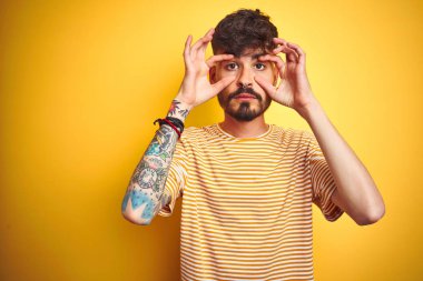 İzole sarı arka plan üzerinde ayakta çizgili t-shirt giyen dövme ile genç adam parmaklar, Uykulu ve sabah yorgunluk için yorgun gözleri açmaya çalışıyorum