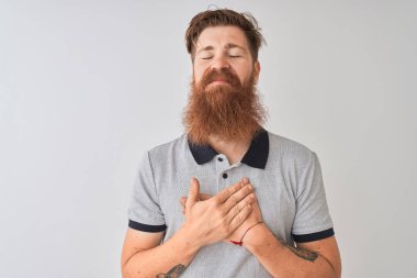 Genç Redhead İrlandalı adam kapalı gözler ve yüz minnettar jest ile göğsüne eller ile gülümseyen izole beyaz arka plan üzerinde ayakta gri Polo giyiyor. Sağlık konsepti.
