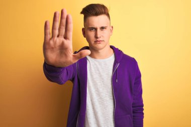 Mor sweatshirt giyen genç yakışıklı adam izole sarı arka plan üzerinde duran avuç içi ile şarkı durdurmak yapıyor. Yüzünde olumsuz ve ciddi bir jest olan uyarı ifadesi.