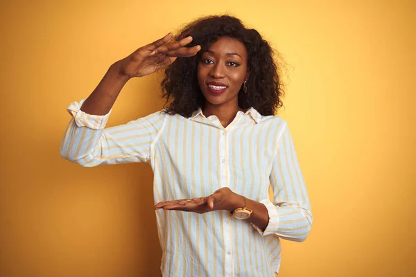 African american woman wearing striped shirt standing over isolated yellow background gesturing with hands showing big and large size sign, measure symbol. Smiling looking at the camera. Measuring concept.