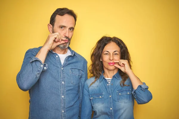Güzel orta yaş çift birlikte izole sarı arka plan ağız üzerinde ayakta ve dudaklar parmaklar ile fermuar olarak kapatıldı. Gizli ve sessiz, tabu konuşan