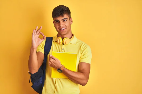 インドの学生の男は 指でOkサインをやって孤立した黄色の背景の上にバックパックヘッドフォンノートブックを身に着けている 優れたシンボル — ストック写真