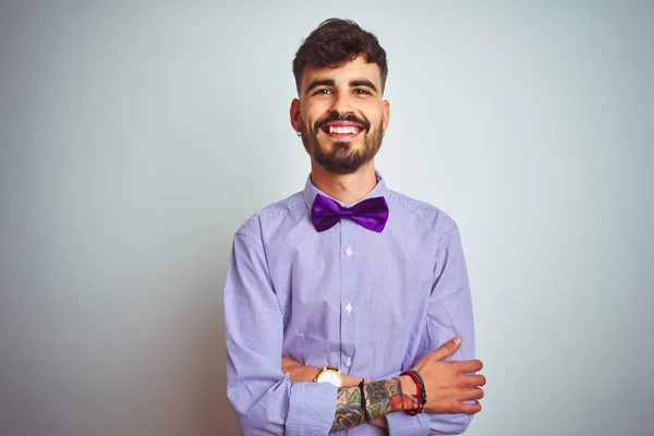 Young man with tattoo wearing purple shirt and bow tie over isolated white background happy face smiling with crossed arms looking at the camera. Positive person.