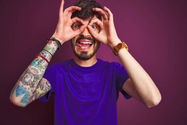 Dövme ile genç adam izole mor arka plan üzerinde t-shirt ayakta giymek dürbün yapışan gibi ok jest yapıyor, gözleri parmaklarla bakıyor. Çılgın ifade.