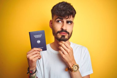 Dövme ile genç adam izole sarı arka plan üzerinde Alman Almanya pasaport giyen soru hakkında düşünme ciddi yüz, çok karışık fikir