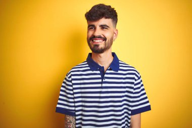 Dövme ile genç adam izole sarı arka plan üzerinde yüzünde gülümseme ile yan bakıyor çizgili polo ayakta giyen, doğal ifade. Güven gülüyor.