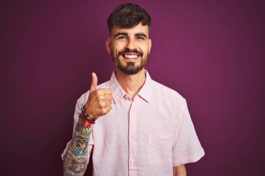 Dövme ile genç adam yaz pembe gömlek ayakta izole mor arka planda duran mutlu başparmak yukarı jest yapıyor ile el. Başarı göstererek kameraya bakarak ifadeyi onaylama.