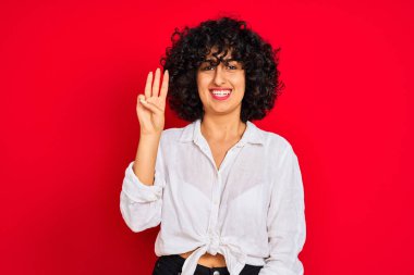İzole kırmızı arka plan üzerinde beyaz rahat gömlek giyen kıvırcık saçlı genç arap kadın gösteren ve emin ve mutlu gülümserken parmaksayısı üç işaret.